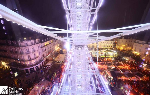 vue immersive et festive du marché de Noël d'Orléans, capturée depuis la grande roue. Elle met en évidence l'illumination intense et féérique des structures blanches de la roue, dominant la place. En contrebas, des chalets en bois, décorés et éclairés, forment un marché animé où les visiteurs se pressent. Autour de la place, des bâtiments élégants illuminés contribuent à l'ambiance chaleureuse et festive. La scène se déroule de nuit, amplifiant l'éclat des lumières et la magie de Noël.