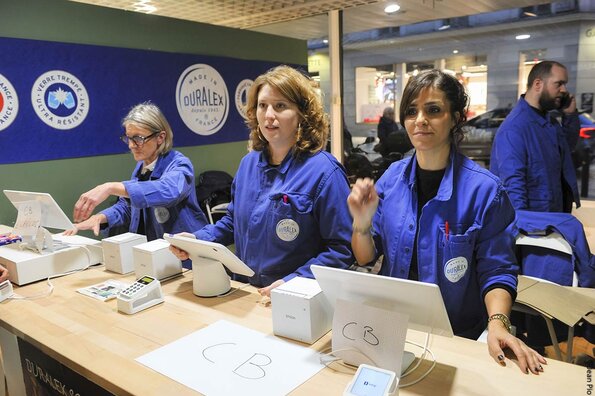 La boutique Duralex d'Orléans. Quatre hôtesses de caisse, portant des blouses de travail bleues avec le logo Duralex, se tiennent derrière un comptoir en bois clair.