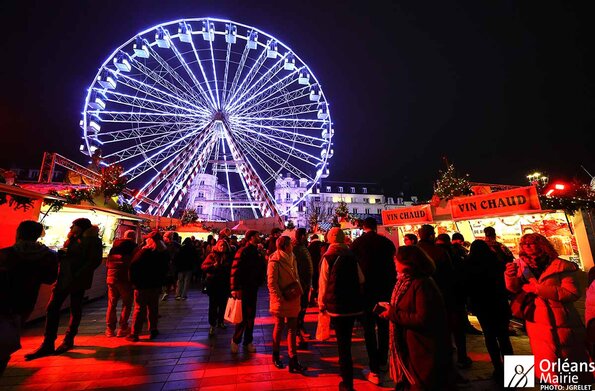 Marché de Noël d'Orléans sur la place du Martroi : Au centre de l'image se dresse une imposante roue panoramique illuminée de lumières bleu vif. Ses grandes structures métalliques s'élèvent dans un ciel sombre, créant un point focal saisissant. Autour de la roue, on peut voir des dizaines de visiteurs rassemblés sur la place. Ils sont emmitouflés dans des manteaux, écharpes et bonnets, profitant de l'ambiance festive du marché de Noël. Des stands de vin chaud et d'autres échoppes traditionnelles ornent la place, leurs auvents rouges et blancs rayés ajoutant une touche de chaleur à la scène. La foule se presse entre ces différents stands, créant une animation palpable. Des guirlandes lumineuses et d'autres décorations de Noël complètent le décor, baignant l'ensemble dans une atmosphère chaleureuse et conviviale. Dans l'ensemble, cette image transmet l'effervescence et la joie d'un marché de Noël urbain en pleine activité, avec sa roue panoramique illuminée comme point focal central.