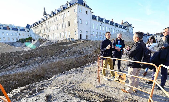 Un groupe de personnes, dont des membres des équipes en charge du projet et des représentants de la presse, visite le chantier du site Porte Madeleine à Orléans le 5 mars. À l’arrière-plan, un bâtiment historique et des tas de terre témoignent des travaux en cours. Une barrière de sécurité est visible au premier plan, délimitant la zone de chantier.