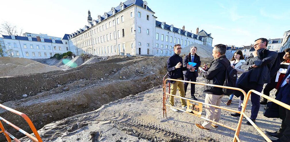 Un groupe de personnes, dont des membres des équipes en charge du projet et des représentants de la presse, visite le chantier du site Porte Madeleine à Orléans le 5 mars. À l’arrière-plan, un bâtiment historique et des tas de terre témoignent des travaux en cours. Une barrière de sécurité est visible au premier plan, délimitant la zone de chantier.