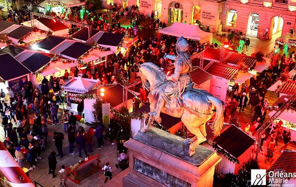 Marché de Noël animé sur la place du Martroi à Orléans, avec en point central la statue équestre de Jeanne d'Arc. On peut voir une foule nombreuse déambulant entre les stands du marché, qui sont illuminés de couleurs chaudes. L'ensemble transmet une ambiance vibrante et conviviale.