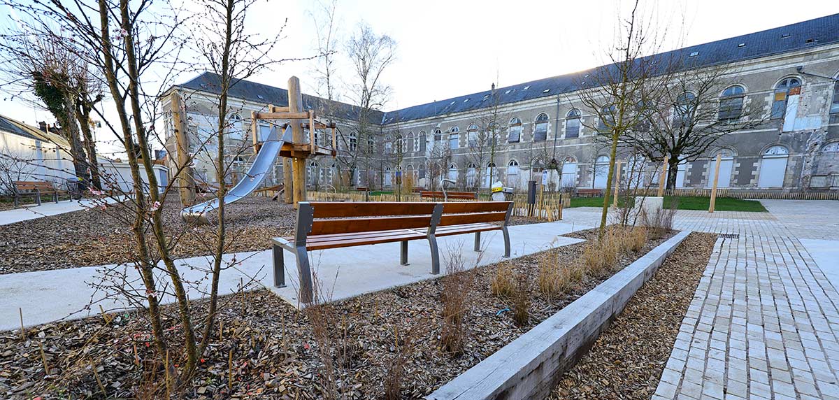 Vue d’un nouveau jardin public à Orléans, aménagé avec des bancs en bois, des allées pavées et des espaces végétalisés. Une aire de jeux en bois avec un toboggan est visible en arrière-plan, entourée d’arbres jeunes et d’arbustes. Le grand bâtiment de l'ancien hopital se dresse à l’arrière du parc, ajoutant une touche patrimoniale à l’ensemble.